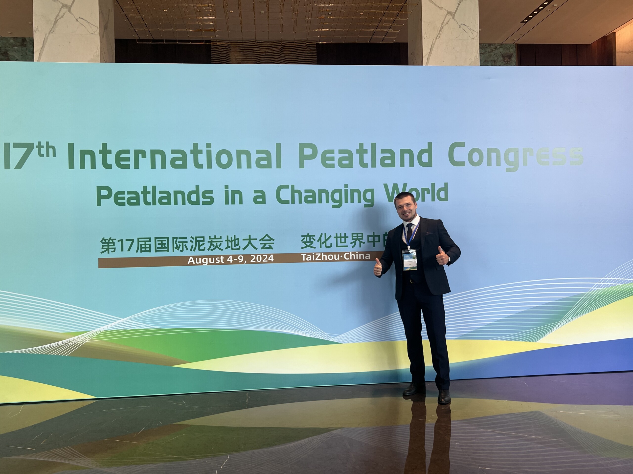 Delegate giving a thumbs-up before the 17th International Peatland Congress banner, Taizhou, China, 2024.