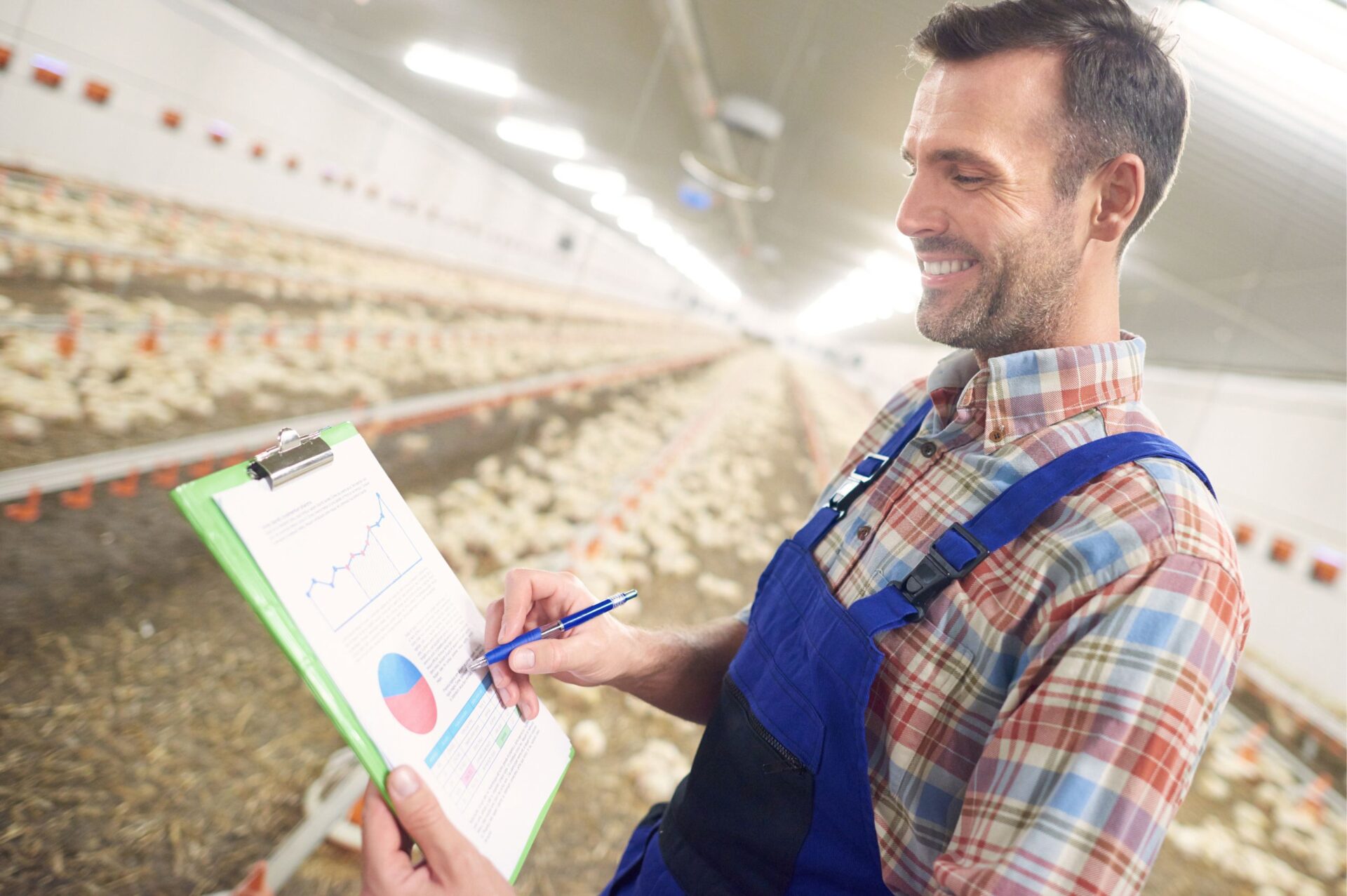 Smiling farmer in blue overalls checking data on a clipboard inside a large poultry farm.