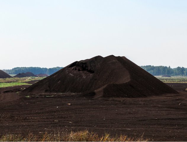 A large pile of harvested low bog peat in an open field, with additional piles and a tree line visible in the background under a clear sky.