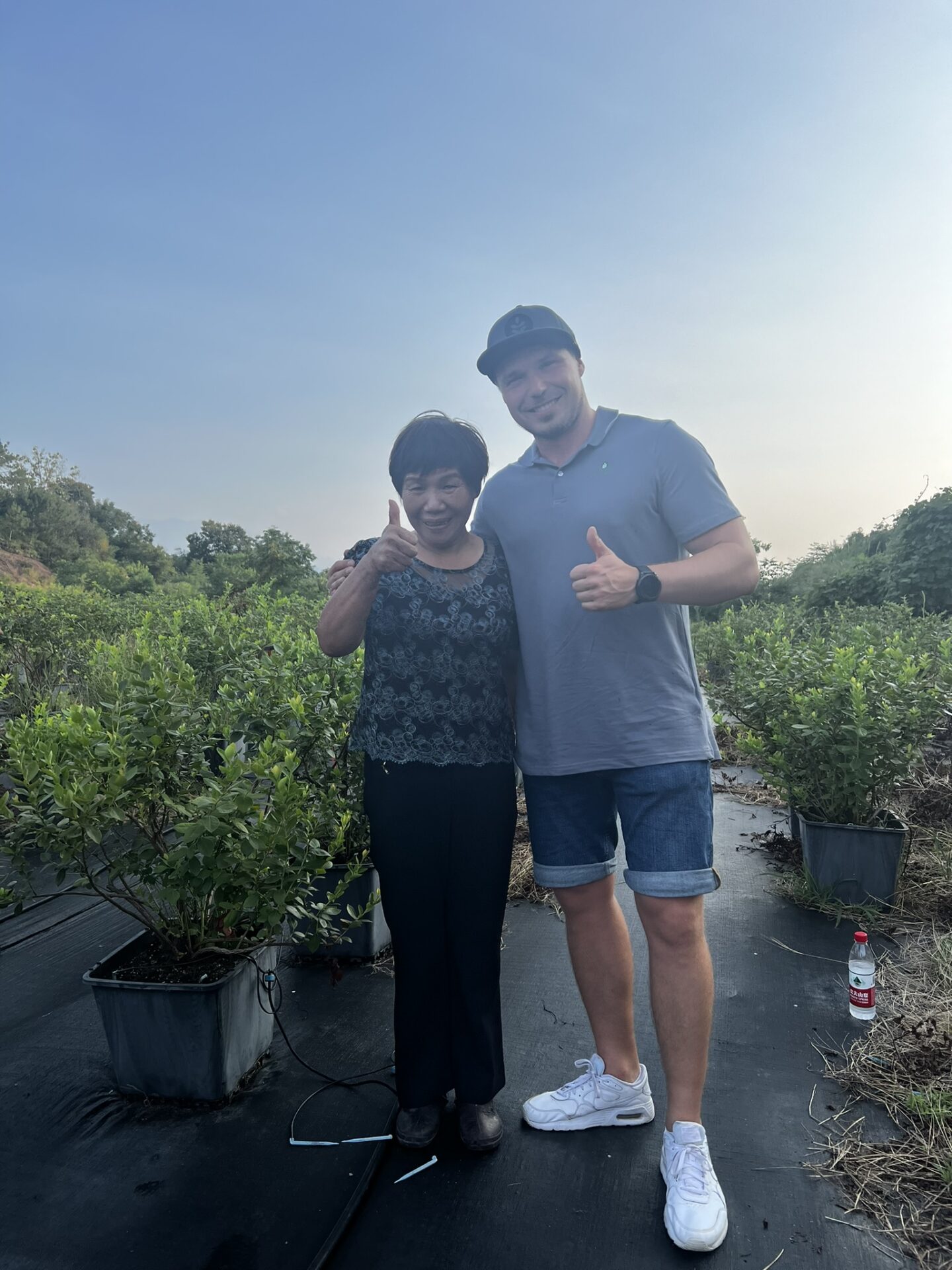 A local grower and a visitor smiling and giving a thumbs-up while standing in a blueberry plantation in China, surrounded by potted plants under a clear blue sky.