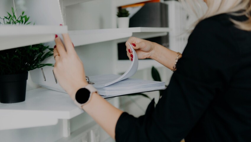 Person reviewing documents in a binder on a shelf, symbolizing organization and paperwork management