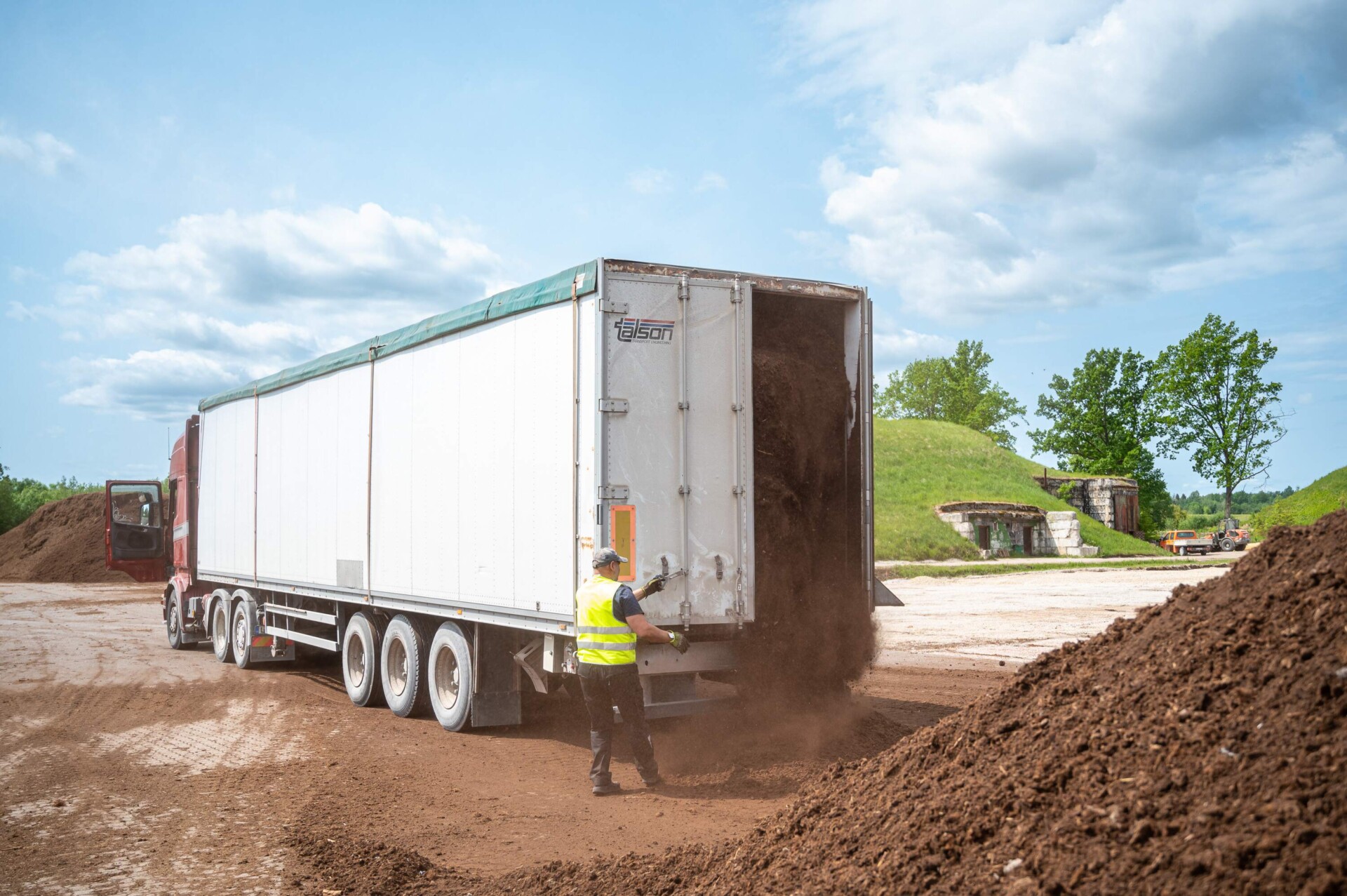 Worker in a yellow ETEPEK vest unloading peat moss from a large truck in an open field.