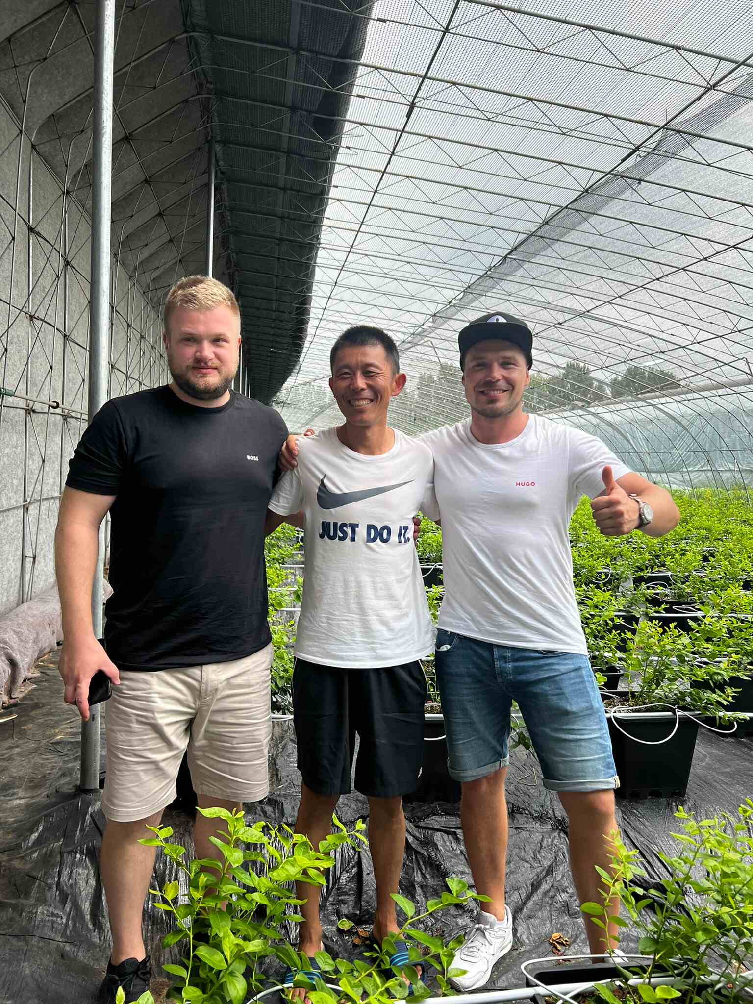 Three men standing together in a greenhouse filled with potted plants, smiling for the camera, with one giving a thumbs-up gesture. The greenhouse structure is visible in the background.