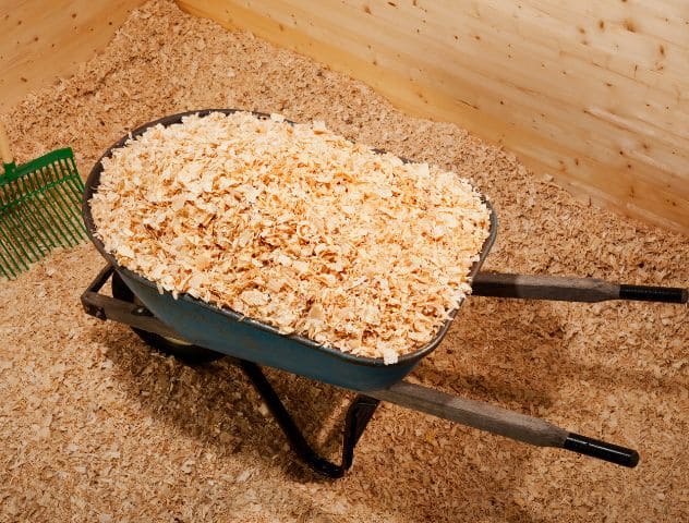 A wheelbarrow filled with fresh wood shavings inside a stable, with a green rake nearby, illustrating the process of maintaining clean and comfortable animal bedding, etepek